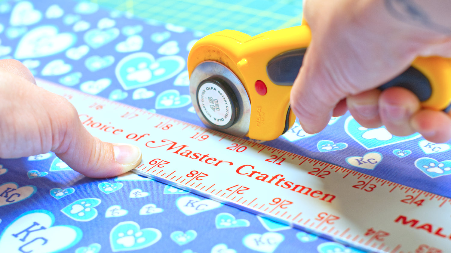 image of SLiK Hound master craftsmen's hands using yellow rotary cuter and grey master craftsmen metal ruler to cut blue KC Hearts Kansas City fabric for cat and dog collars and leashes to an exact specification. All part of the fine tuned process used to make the worlds best dog collars