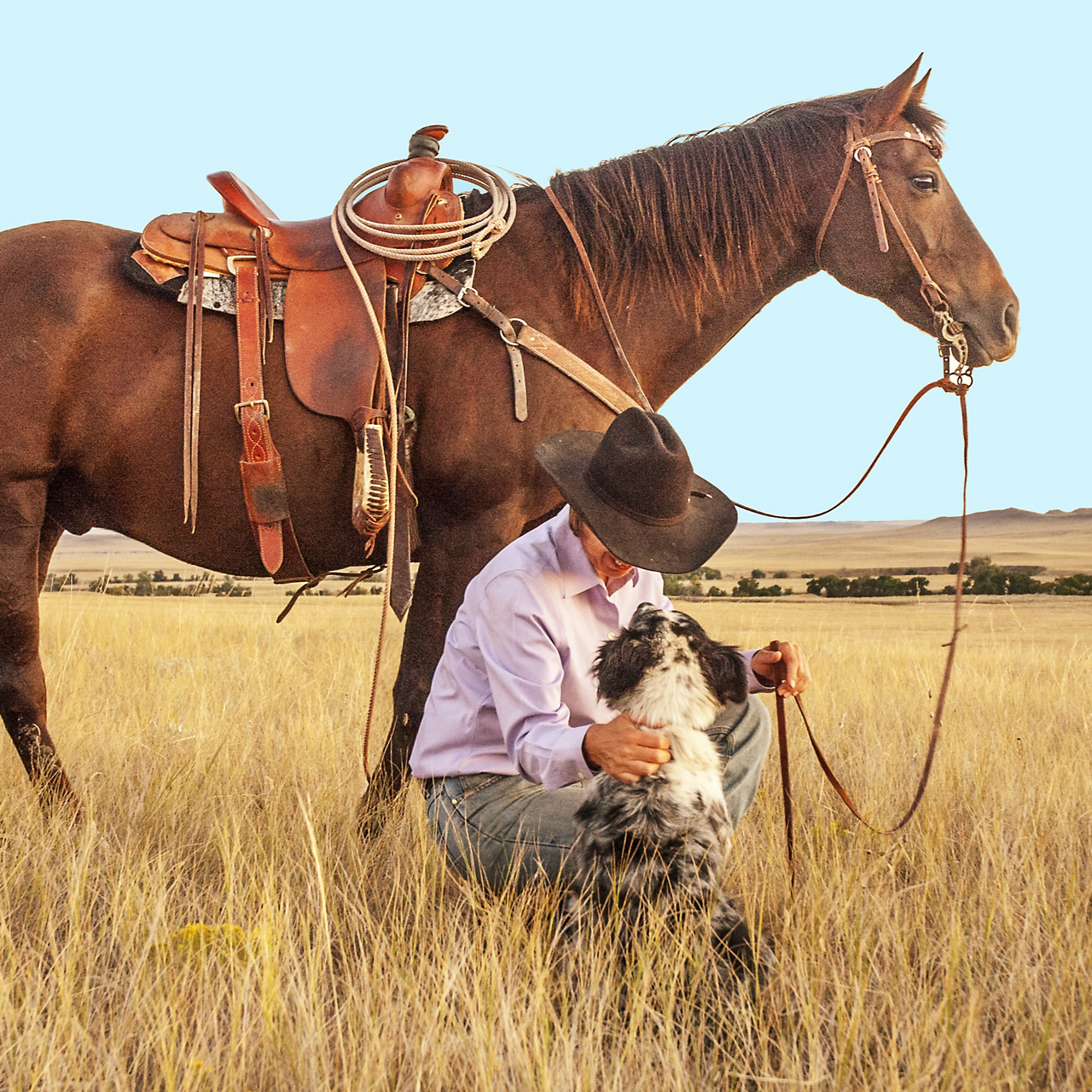 Man in black cowboy hat kneeling next to SLiK Hound western dog while holding the reins of the brown quarter horse behind them. Pictured in knee high tan wheat grass with rolling hills and blue sky over head.