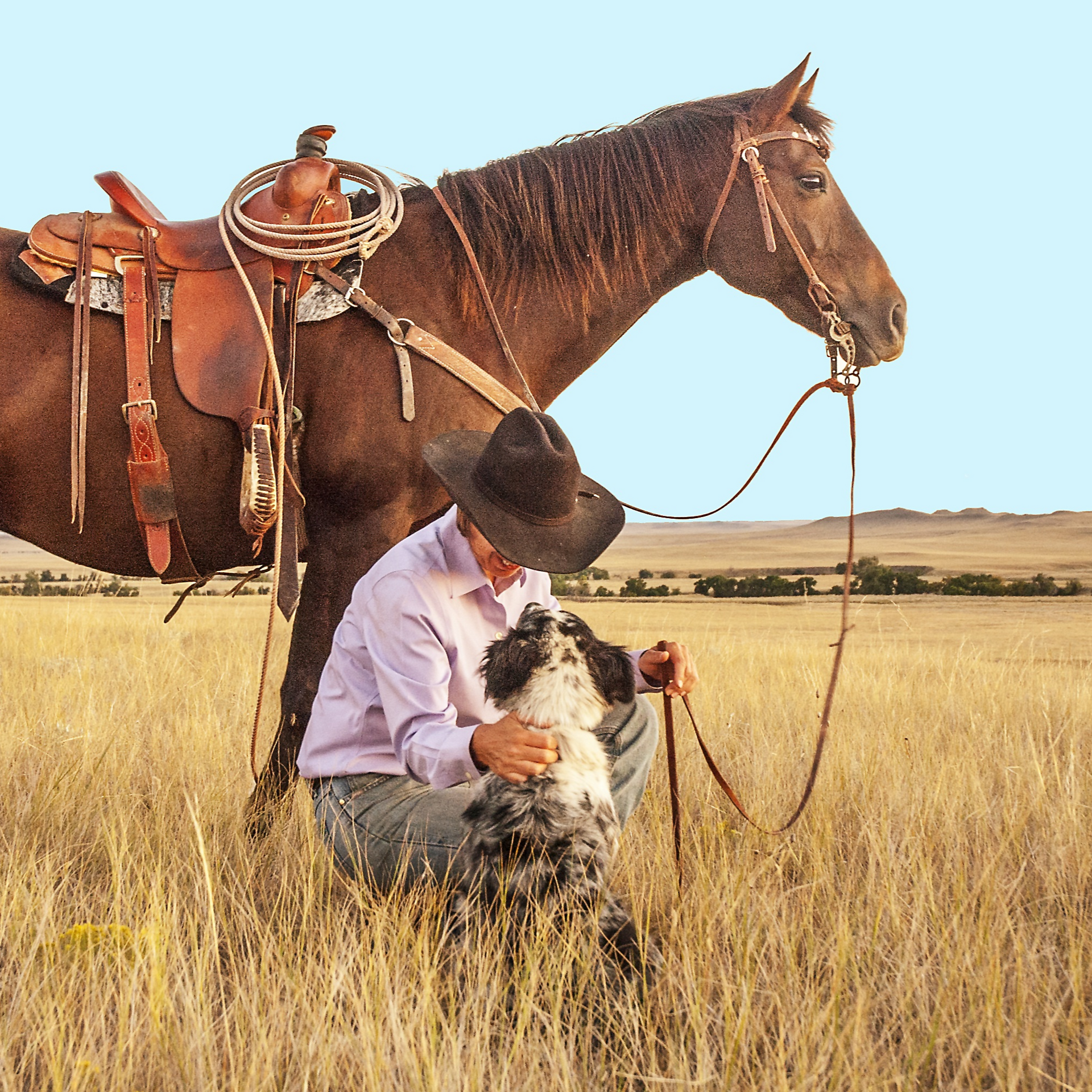 Cowboy leaning next to brown horse with leather saddle. The cowboy is leaned down with their hand over a white and black dog in a field of tan grasses and a blue sky. wearing western themed dog collar by SLiK hound