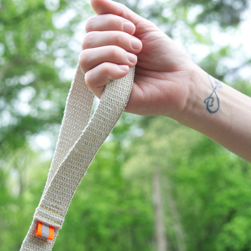 Tan undyed and nature hemp Canva leash handle for dogs with fluorescent orange reflective tabs at all ends, yellow kevlar stitching being held by hand with paw print tattoo wrist against a green forest backdrop.