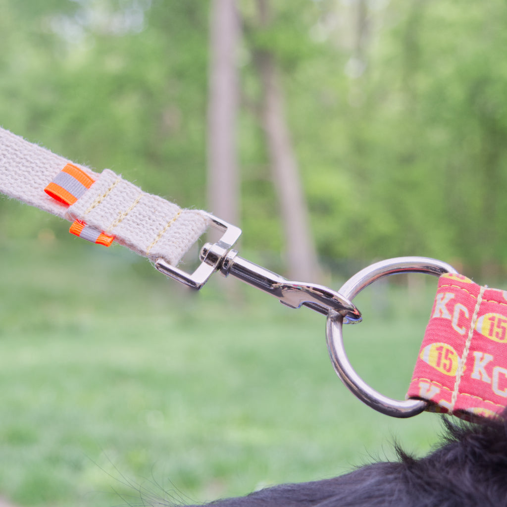 Close up picture  of Tan undyed and nature hemp Canva leash hook for dogs with fluorescent orange reflective tabs at all ends, yellow kevlar stitching and shiny silver metal snap hook fastened to shiny metal D ring of slik hound collar being worn by black dog just out of frame and a blurry forest green backdrop.
