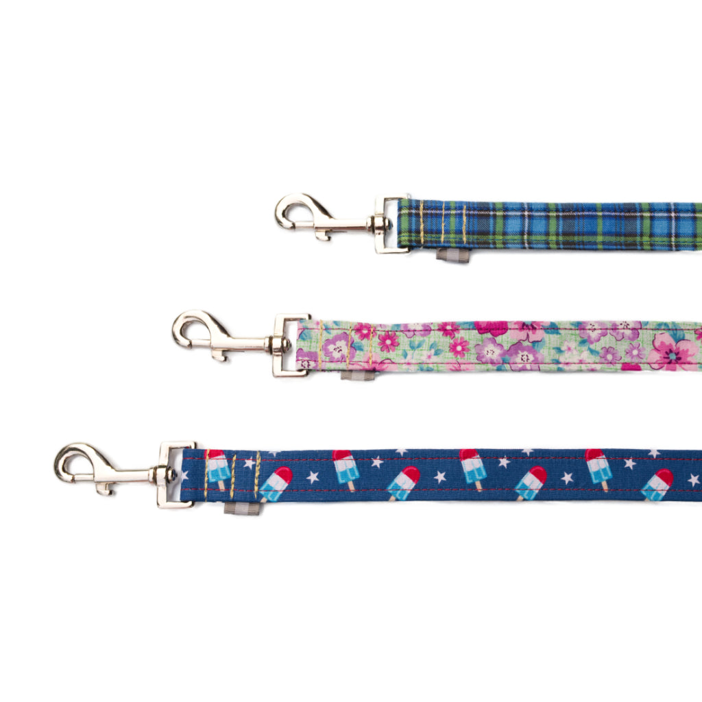 A collection of SLiK Hound reflective dog leashes in various patterns including green blue plaid, pink and purple sea foam floral, and a red, white, and blue bomb pop design each with a metal clasp pictured against a white backdrop.