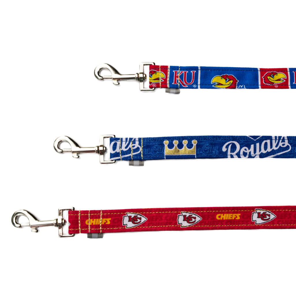 A red kc chiefs NFL dog leash next to a blue kc royals dog leash next to a red and blue Kansas Jayhawks dog leash all with silver quick realize hooks and reflective safety tabs.