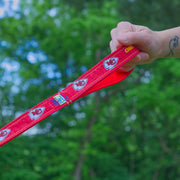 Bright red Kansas City Chiefs leash with arrowhead kc logos being held by hand with dog tattoo wrist with a green forest background.