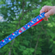 close up image of hand holding the handle of a SLiK Hound reflective leash in red, white, and blue bomb pop pattern with red stitching against a forest backdrop