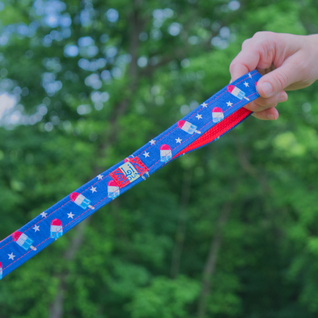 close up image of hand holding the handle of a SLiK Hound reflective leash in red, white, and blue bomb pop pattern with red stitching against a forest backdrop